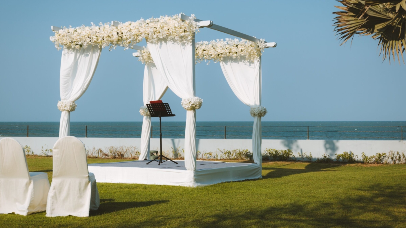 A wedding pavilion adorned by white curtains and artificial flowers set for an outdoor garden wedding by the sea